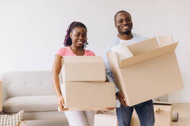 Smiling African American couple carrying cardboard boxes while moving into their new home, embracing the excitement of fresh beginnings