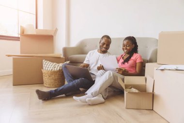 Smiling african american couple sitting on floor among cardboard boxes, using laptop and reading documents, organizing move to new home