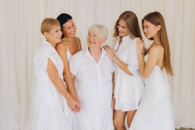 Family members of different generations wearing white dresses are smiling and embracing, celebrating their bond