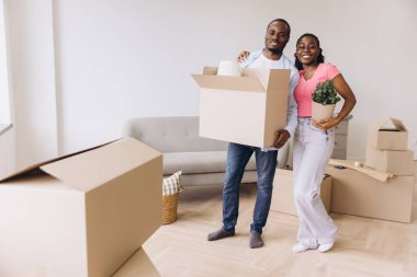 Young smiling african american couple carrying cardboard boxes and a plant into their new apartment