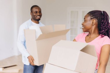 Young African American couple smiling while carrying cardboard boxes, joyfully moving into their new house and starting a bright future together