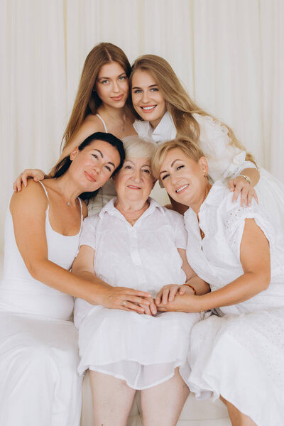 Family portrait of women of different generations hugging their grandmother, wearing white dresses and sitting on a white sofa