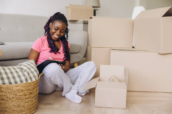 Smiling african american woman checking boxes while moving into new home, creating inventory list and organizing belongings