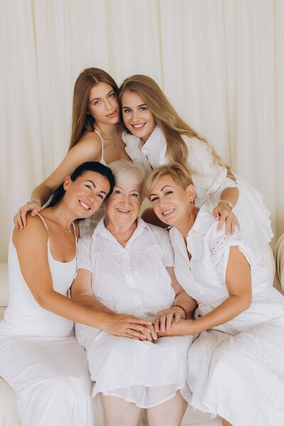 Five cheerful women of different ages wearing white dresses are hugging each other, celebrating their family bond