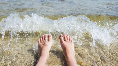 Kadın bacakları sahilde, güneşin altında bir dalga arkaplanda.