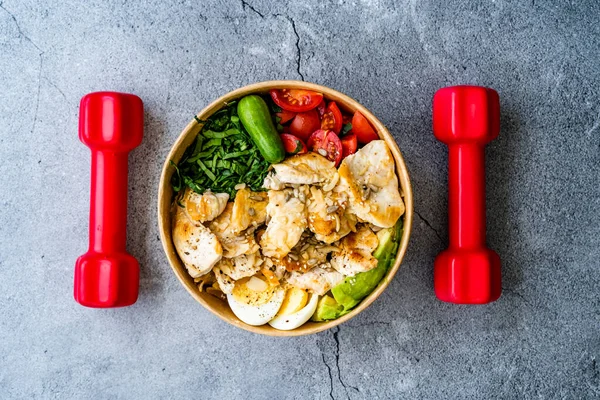 Healthy Protein Fitness Food Chicken, Egg, Spinach, Avocado and Cucumber with Fitness Equipment Dumbbells for Athlete in Plastic Bowl. Ready to Eat.