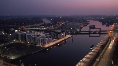 Geceleri Polonya 'daki Szczecin şehir manzarasının insansız hava aracı panoraması. Manzarada Oder nehri üzerindeki aydınlık köprüler, modern deniz manzarası mimarisi ve arka planda parlayan şehir ışıkları yer alıyor. Yollarda araba trafiği ve
