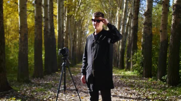 Фотограф стоит рядом с треногой и надевает очки в парке — стоковое видео
