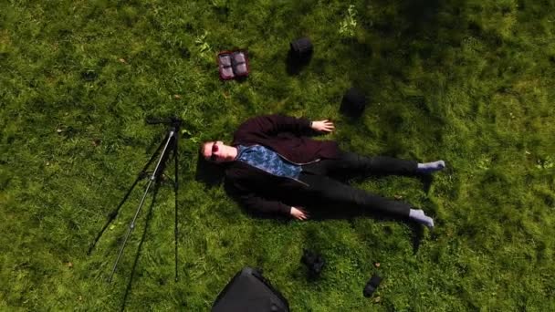 Air shoot, the guy is lying on the green grass with photo equipment a dog runs up to him — Stock Video