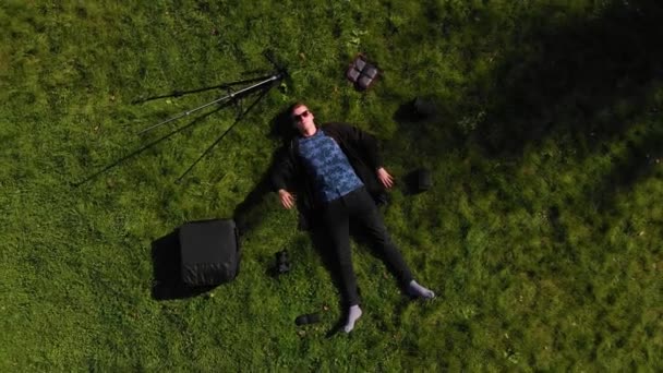 A person lies surrounded by equipment on a sunny green glade of the park, the camera pulls off and the person puts his hand under his head — Stock Video