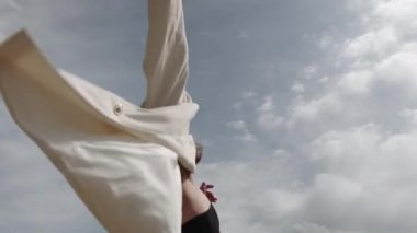 Esmer kız, beyaz bir kostüm ve siyah bir bluzla gökyüzüne karşı şehvetle dans ediyor.