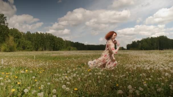 Menina ruiva encaracolado em um vestido florido dança e, em seguida, cai na grama e dentes de leão em um fundo de floresta e céu à luz do dia — Vídeo de Stock