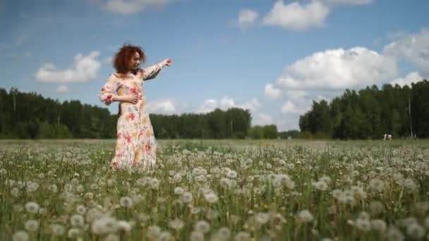 Menina encaracolada ruiva em um vestido florido dança em um fundo de floresta e céu à luz do dia — Vídeo de Stock