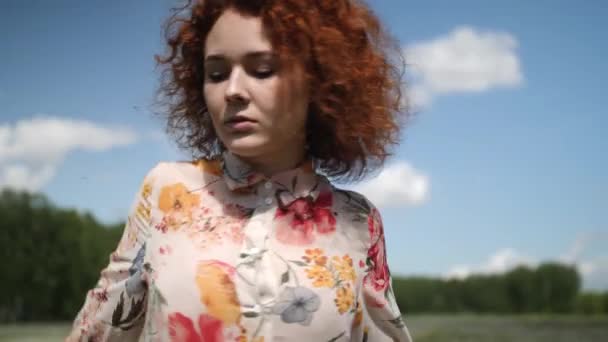Rothaarige, lockige Mädchen in einem geblümten Kleid tanzt auf einem Hintergrund aus Wald und Himmel bei Tageslicht — Stockvideo
