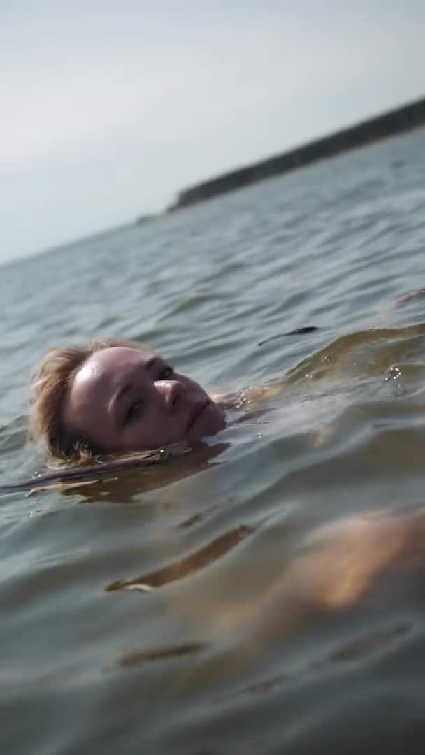 Ξανθιά κοπέλα κολυμπάει στο νερό και κοιτάζει την κάμερα. — Αρχείο Βίντεο