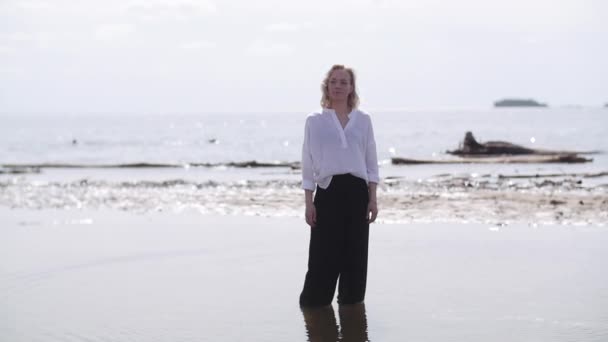 Ξανθιά κοπέλα με άσπρα και μαύρα ρούχα στέκεται στο νερό κοιτάζοντας την κάμερα και χαμογελώντας στο φόντο της ακτής και του νερού — Αρχείο Βίντεο