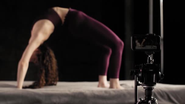 A yoga teacher teaches exercises through an online conference. The girl practices yoga on the bed, on a black background — Stock Video