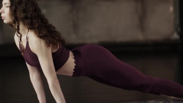 Kudrnatá holka s dlouhými vlasy dělá jógu na podložce. Mladá dívka v burgundském oblečení sportu — Stock video