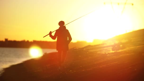 Seorang nelayan dengan pancing berjalan di sepanjang pantai ke kamera. Shooting in gold time — Stok Video