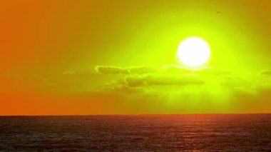 Denizin üzerinde gün batımı. Güneş kısmen bulutlar tarafından gizlenir, gökyüzünde altın ve turuncu renkler bırakır ve okyanus dalgalarını yansıtır..