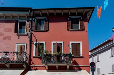 İtalya 'nın Desenzano del Garda şehrinde süslü saksı bitkileriyle süslenmiş ahşap panjurları ve işlenmiş demir balkonları olan parlak kırmızı renkli bir bina. Dekoratif kelebekler büyüleyici Akdeniz caddesinde asılı.