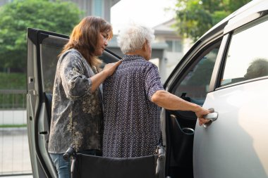 Bakıcı, Asyalı yaşlı kadın maluliyet hastasının arabasına binmesine yardım ediyor..