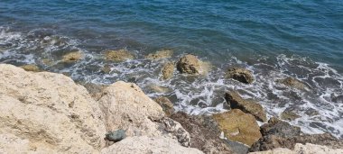 Kıbrıs Rum Kesimi 'nin Akdeniz kıyısındaki engebeli kayaların kıyıya vuran hafif dalgalarla yakın çekim görüntüsü. Açık mavi su ve doğal dokular, saf güzelliğin altını çiziyor.