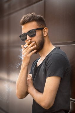 Bir genç yakışıklı beyaz sakallı adam güneş gözlüğü ve siyah tişört ilkbaharda sokakta sigara içiyor. Yakın çekim.