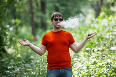 Genç bir beyaz sakallı kırmızı t-shirt ve güneş gözlüğü vaping ormanın içinde elektronik sigara yaz aylarında adamdır. Yakın çekim.