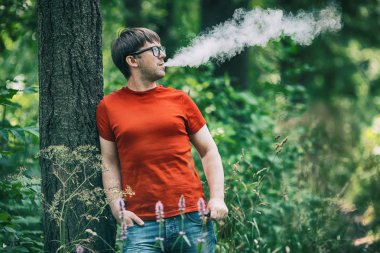 Genç bir beyaz sakallı kırmızı t-shirt ve gözlük vaping ormanın içinde elektronik sigara yaz aylarında adamdır. Yakın çekim.