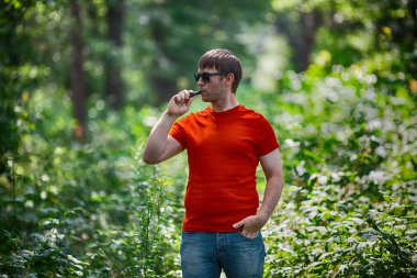 Genç bir beyaz sakallı kırmızı t-shirt ve güneş gözlüğü vaping ormanın içinde elektronik sigara yaz aylarında adamdır. Yakın çekim.
