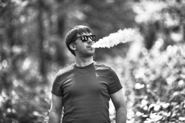 Genç bir beyaz sakallı t-shirt ve güneş gözlüğü vaping ormanın içinde elektronik sigara yaz aylarında adamdır. Yakın çekim. Siyah ve beyaz.