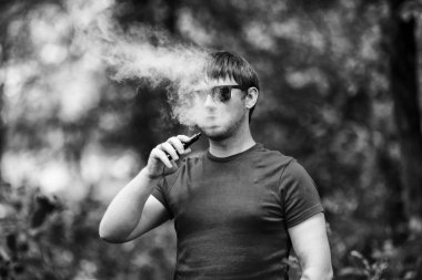 Genç bir beyaz sakallı t-shirt ve güneş gözlüğü vaping ormanın içinde elektronik sigara yaz aylarında adamdır. Yakın çekim. Siyah ve beyaz.