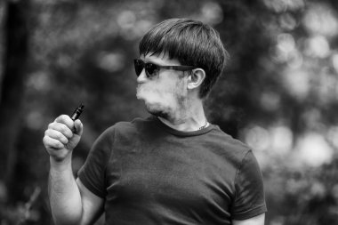 Genç bir beyaz sakallı t-shirt ve güneş gözlüğü vaping ormanın içinde elektronik sigara yaz aylarında adamdır. Yakın çekim. Siyah ve beyaz.