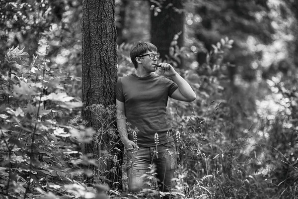 一个年轻的白胡子男子穿着 T恤衫 戴着眼镜 夏天在森林里 Vaping 一支电子香烟 关门了黑白相间 — 图库照片