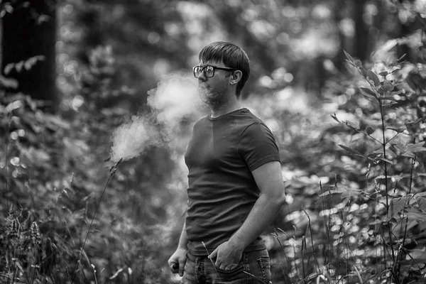 A young white bearded man in t-shirt and in glasses is vaping an electronic cigarette in the forest in the summer. Close up. Black and white.