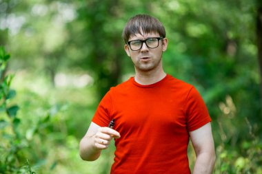 Genç bir beyaz sakallı kırmızı t-shirt ve gözlük vaping ormanın içinde elektronik sigara yaz aylarında adamdır. Yakın çekim.