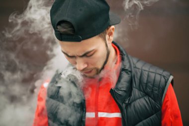Vape genç. Yakışıklı genç bir beyaz adam siyah kap vaping sonbahar modern arka planda karşısında elektronik sigara portresi.
