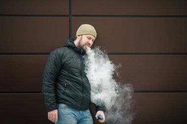 Vape adam. Genç sakallı kap beyaz adam ve siyah ceket bir elektronik sigara ve buhar karşısında modern kahverengi zemin kapalı icar. Yaşam tarzı.