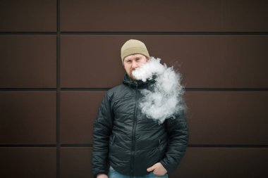 Vape adam. Genç sakallı kap beyaz adam ve siyah ceket bir elektronik sigara ve buhar karşısında modern kahverengi zemin kapalı icar. Yaşam tarzı.