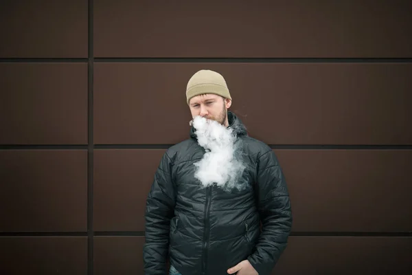 Vape man. Young bearded white guy in the cap and black jacket is smoking an electronic cigarette and letting off the steam opposite modern brown background. Lifestyle.