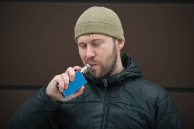 Vape adam. Genç sakallı kap beyaz adam ve siyah ceket modern kahverengi zemin karşısında elektronik sigara sigara içiyor. Bir yaşam tarzı. Yakın çekim.