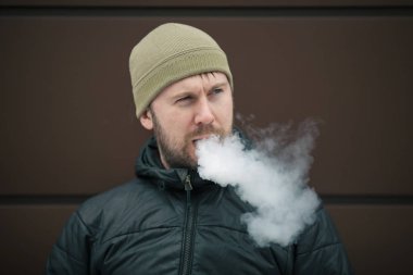 Vape adam. Genç sakallı kap beyaz adam ve siyah ceket bir elektronik sigara ve buhar karşısında modern kahverengi zemin kapalı icar. Bir yaşam tarzı. Yakın çekim.