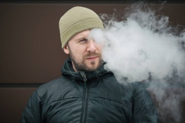 Vape adam. Genç sakallı kap beyaz adam ve siyah ceket bir elektronik sigara ve buhar karşısında modern kahverengi zemin kapalı icar. Bir yaşam tarzı. Yakın çekim.