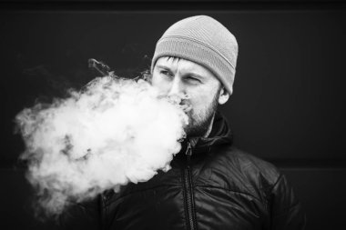 Vape adam. Genç sakallı beyaz şapkalı adam bir elektronik sigara ve buhar icar. Kötü alışkanlık. Siyah ve beyaz. Yakın çekim.