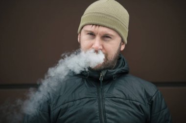 Vape adam. Genç sakallı kap beyaz adam ve siyah ceket bir elektronik sigara ve buhar karşısında modern kahverengi zemin kapalı icar. Bir yaşam tarzı. Yakın çekim.