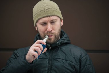 Vape adam. Genç sakallı kap beyaz adam ve siyah ceket modern kahverengi zemin karşısında elektronik sigara sigara içiyor. Bir yaşam tarzı. Yakın çekim.