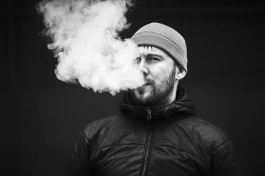 Vape adam. Genç sakallı beyaz şapkalı adam bir elektronik sigara ve buhar icar. Kötü alışkanlık. Siyah ve beyaz. Yakın çekim.