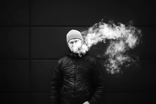 Vape man. Young bearded white guy in the cap is smoking an electronic cigarette and letting off the steam. Bad habit. Black and white.