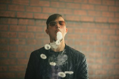 Vaping genç. Genç yakışıklı sakallı beyaz adam bir siyah ceket ve güneş gözlüğü elektronik sigara sigara ve buhar vape çubuğunda puffs kapalı icar. Kötü alışkanlık.